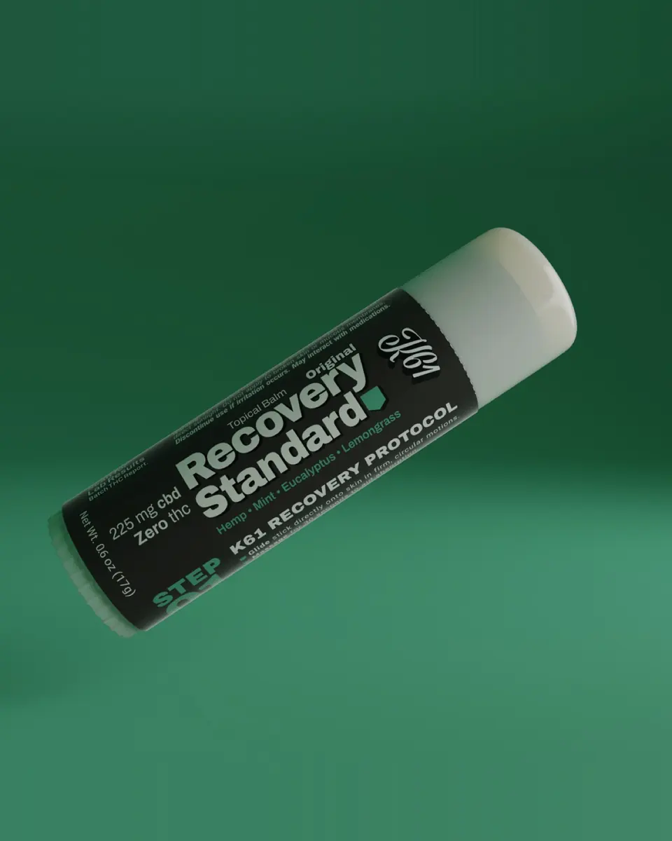 K61 Recovery Standard CBD balm stick with its label facing forward on a green background